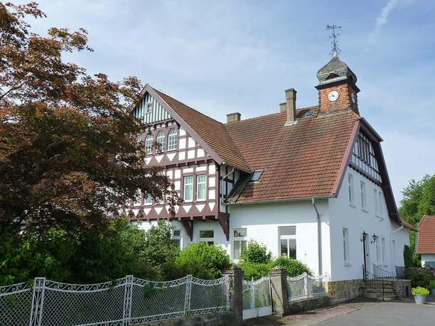 Sattelmeierhof Ringsthof Fachwerkhaus mit roten Ziegeldach, Turmuhr, umgeben von grünen Büschen und einem malerischen Zaun.
