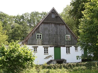 Alte Wassermühle in ländlicher Umgebung, halb Fachwerk, halb gemauert, flankiert von Bäumen.