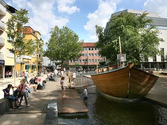 Großer Platz mit Holzschiff-Spielplatz, umgeben von Geschäften und Bäumen, bei sonnigem Wetter.