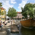 Linnenbauerplatz Großer Platz mit Holzschiff-Spielplatz, umgeben von Geschäften und Bäumen, bei sonnigem Wetter.