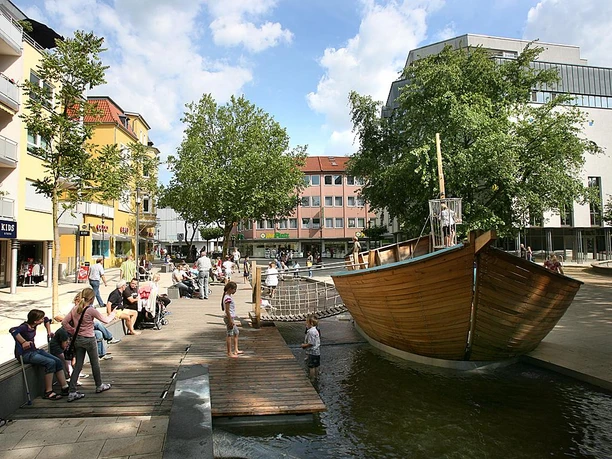 Linnenbauerplatz Großer Platz mit Holzschiff-Spielplatz, umgeben von Geschäften und Bäumen, bei sonnigem Wetter.