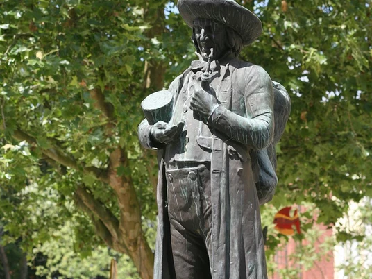 Bronzestatue eines Mannes mit Hut, Pfeife und Bündel. Hintergrund aus Laubbäumen im Sommer.