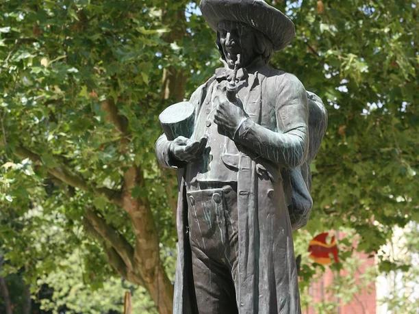 Linnenbauerplatz Denkmal Bronzestatue eines Mannes mit Hut, Pfeife und Bündel. Hintergrund aus Laubbäumen im Sommer.