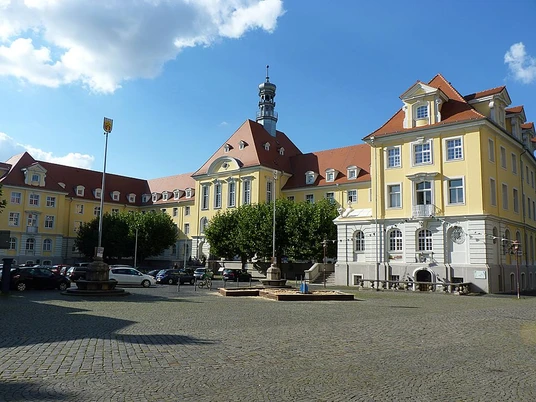 Rathaus, Markthalle und Kuratorium Historisches Gebäudeensemble mit gelben Fassaden und rotem Dach, umgeben von gepflastertem Platz.