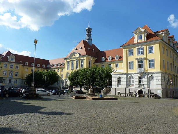 Rathaus, Markthalle und Kuratorium Historisches Gebäudeensemble mit gelben Fassaden und rotem Dach, umgeben von gepflastertem Platz.