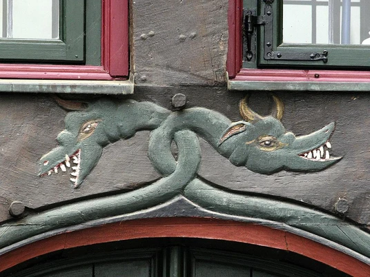 Verzierter Holzgiebel mit zwei abstrahierten Drachenköpfen, vor einem Gebäude aus Fachwerk.