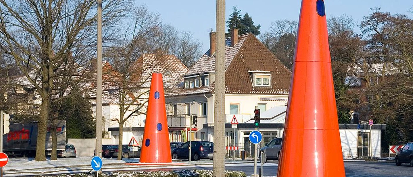 Safety Cones Große rote Kegel auf einem schneebedeckten Kreisverkehr in urbaner Umgebung mit Häusern im Hintergrund.