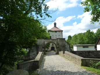 Zufahrt zum historischen Gut Bustedt über eine Steinbrücke, umgeben von grünen Bäumen und blauer Himmel.