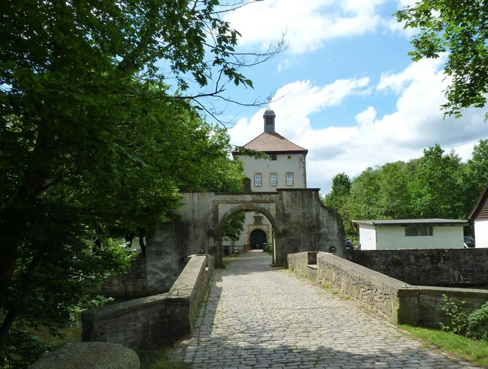 Zufahrt zum historischen Gut Bustedt über eine Steinbrücke, umgeben von grünen Bäumen und blauer Himmel.