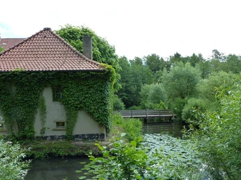Ein von Efeu bewachsenes Gebäude an einem Gewässer mit einer kleinen hölzernen Brücke im Grünen.