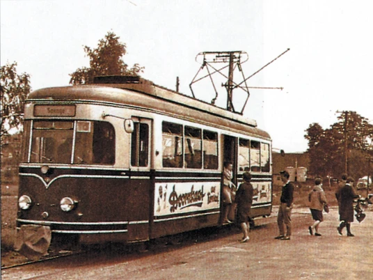 Ehemalige Kleinbahn Historische Straßenbahn mit Werbung, mehrere Menschen steigen ein, umgeben von Bäumen.