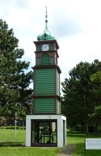 Grüner Holzturm mit Uhr und Satteldach, umgeben von grünen Bäumen und einer Rasenfläche.