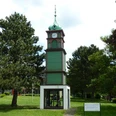 MeyLip-Turm Grüner Holzturm mit Uhr und Satteldach, umgeben von grünen Bäumen und einer Rasenfläche.