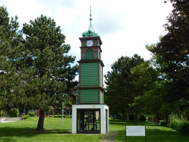 MeyLip-Turm Grüner Holzturm mit Uhr und Satteldach, umgeben von grünen Bäumen und einer Rasenfläche.