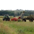 Ein Traktor zieht eine Heupresse über ein Feld, während ein großer Anhänger mit gepresstem Heu beladen wird.