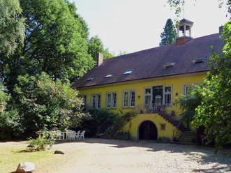 Gelbes Herrenhaus mit begrüntem Treppenaufgang, umgeben von Bäumen, rechts weiße Gartenmöbel.