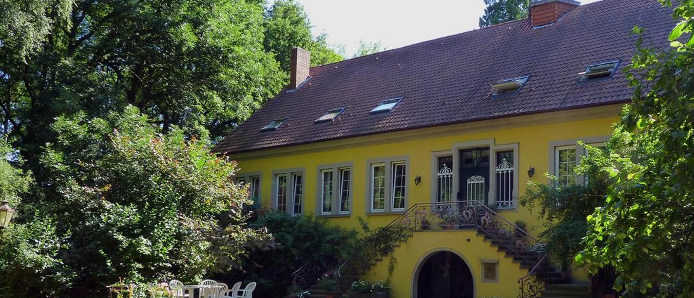 Gelbes Herrenhaus mit begrüntem Treppenaufgang, umgeben von Bäumen, rechts weiße Gartenmöbel.