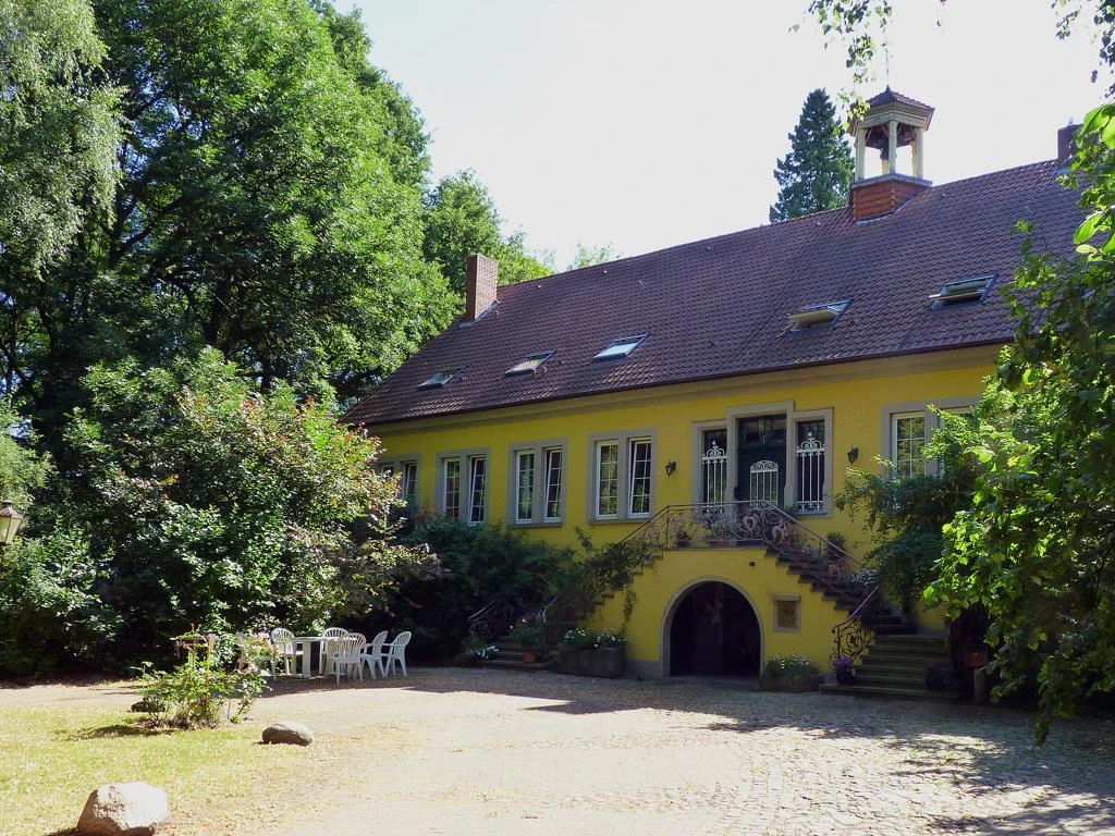 Haus Beck Gelbes Herrenhaus mit begrüntem Treppenaufgang, umgeben von Bäumen, rechts weiße Gartenmöbel.