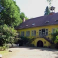 Haus Beck Gelbes Herrenhaus mit begrüntem Treppenaufgang, umgeben von Bäumen, rechts weiße Gartenmöbel.