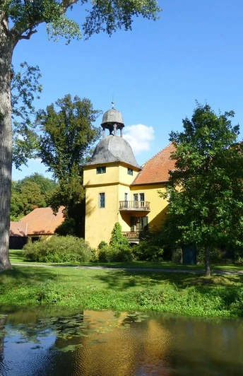 Schloss Gut Böckel mit gelben Wänden und einem Turm inmitten grüner Bäume reflektiert im Wasser.