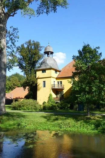 Schloss Gut Böckel mit gelben Wänden und einem Turm inmitten grüner Bäume reflektiert im Wasser.