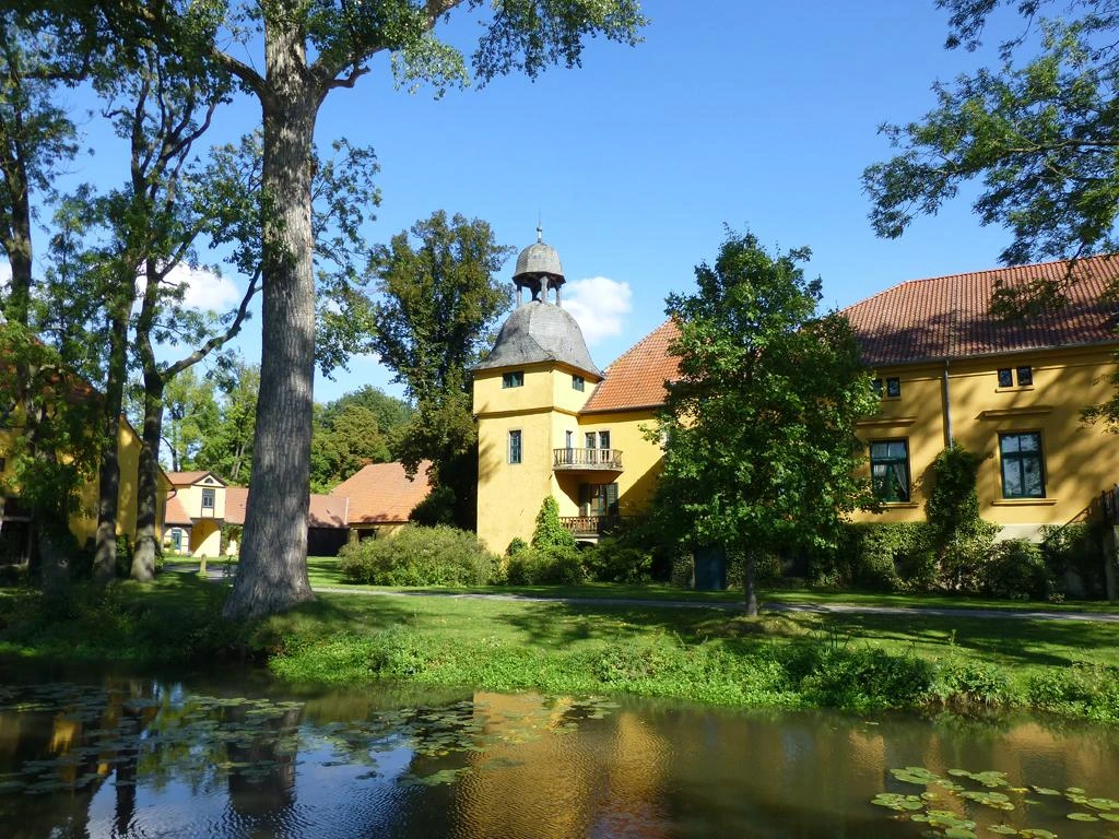Gut Böckel Schloss Gut Böckel mit gelben Wänden und einem Turm inmitten grüner Bäume reflektiert im Wasser.