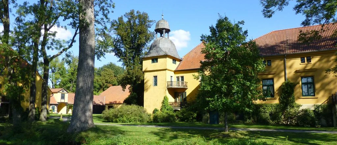Gut Böckel Schloss Gut Böckel mit gelben Wänden und einem Turm inmitten grüner Bäume reflektiert im Wasser.