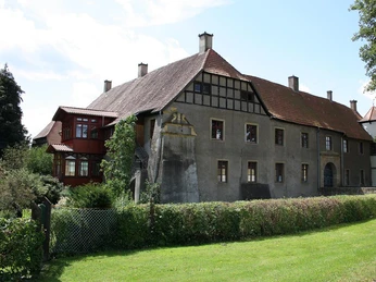 Historisches Gebäude mit Fachwerkelementen in einer grünen Umgebung, umgeben von Bäumen und Sträuchern.