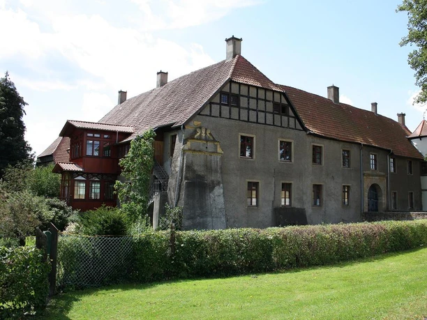 Historisches Gebäude mit Fachwerkelementen in einer grünen Umgebung, umgeben von Bäumen und Sträuchern.