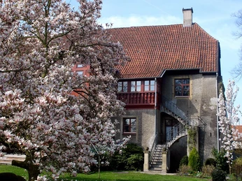 Rotes Backsteinhaus mit markantem Treppeneingang, umgeben von blühenden Magnolienbäumen im Vordergrund.