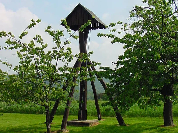 Holzglockenturm auf einer Wiese, umgeben von Kirschbäumen, vor einer bewaldeten Hügelkulisse.