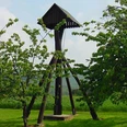 Bauernschaftsglocke Stukenhöfen Holzglockenturm auf einer Wiese, umgeben von Kirschbäumen, vor einer bewaldeten Hügelkulisse.