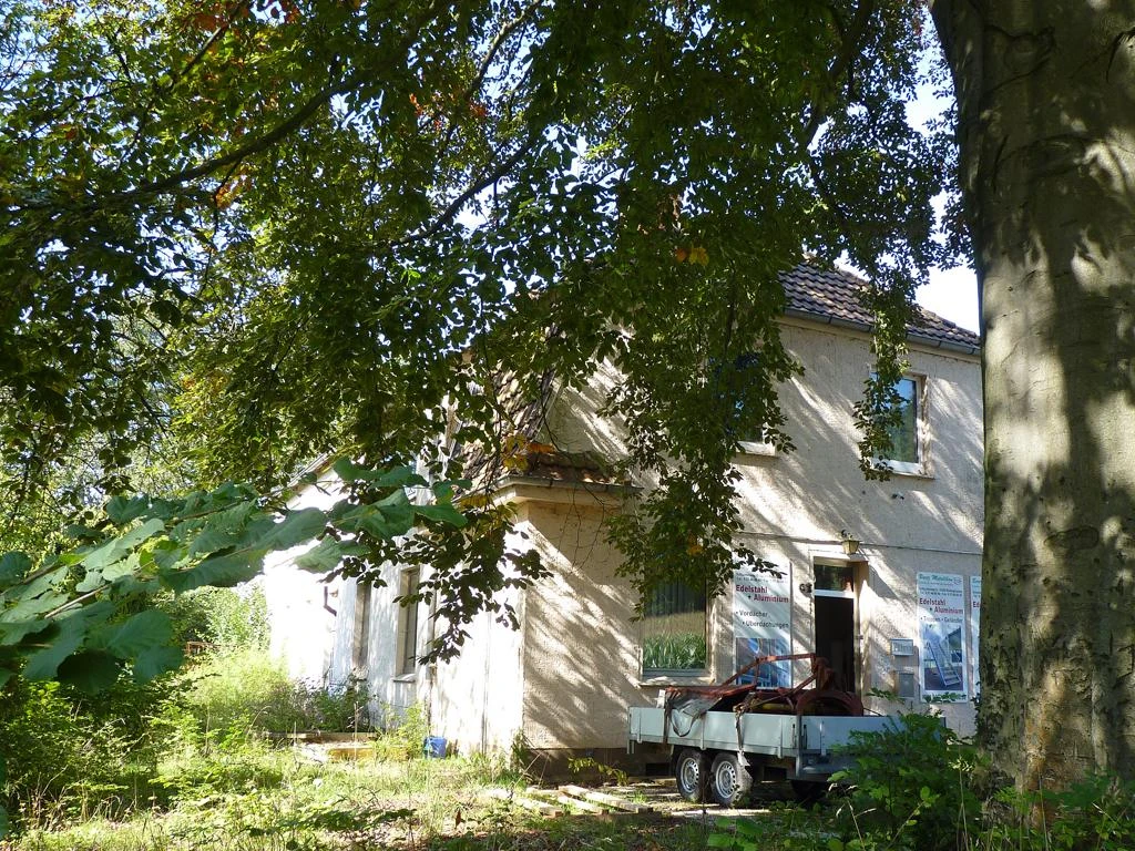 Zweistöckiges, verlassenes Gebäude umgeben von üppigem Grün, im Vordergrund ein geparkter Anhänger.