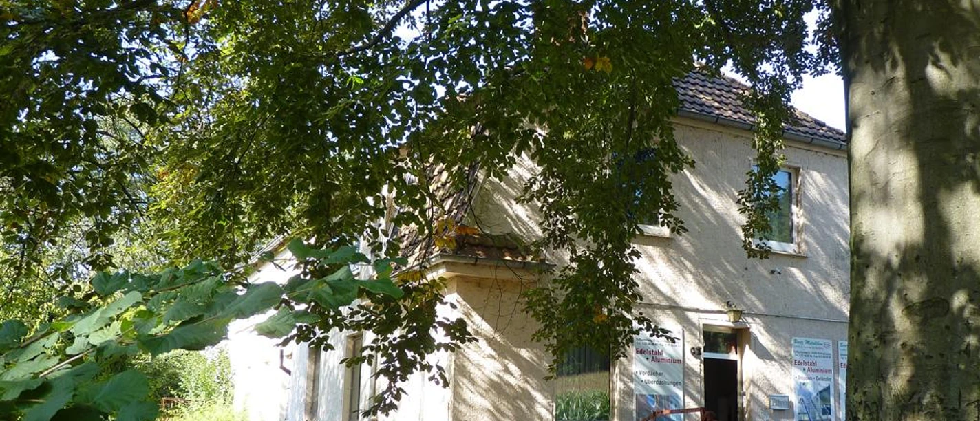 Ehemalige Abdeckerei Zweistöckiges, verlassenes Gebäude umgeben von üppigem Grün, im Vordergrund ein geparkter Anhänger.