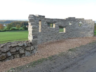 Steinerne Mauer mit Fensteröffnungen auf einem Naturerlebnispfad, umgeben von grünen Feldern.