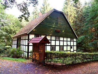 Fachwerkhaus im Grünen, umgeben von Bäumen, mit Schindeldach und dekorativem Holzportal im Vordergrund.