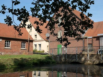 Historische Martmühle mit roten Ziegeldächern, von Bäumen umrahmt, an einem kleinen Gewässer.