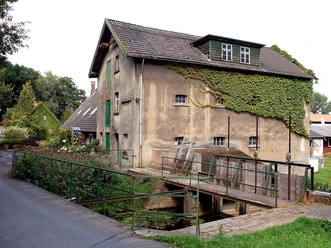 Historische Wassermühle aus Stein mit Efeu bewachsen, neben einer schmalen Brücke und Straße.