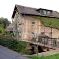 Historische Wassermühle aus Stein mit Efeu bewachsen, neben einer schmalen Brücke und Straße.