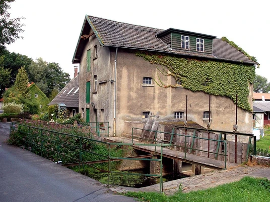 Niedermühle Historische Wassermühle aus Stein mit Efeu bewachsen, neben einer schmalen Brücke und Straße.