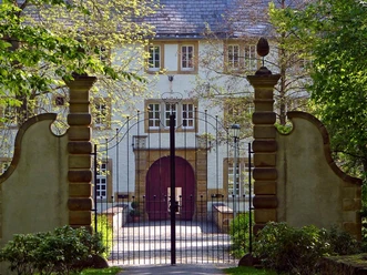 Historisches Schlossgebäude umgeben von schmiedeeisernem Tor und Natur, bei Tageslicht erleuchtet.