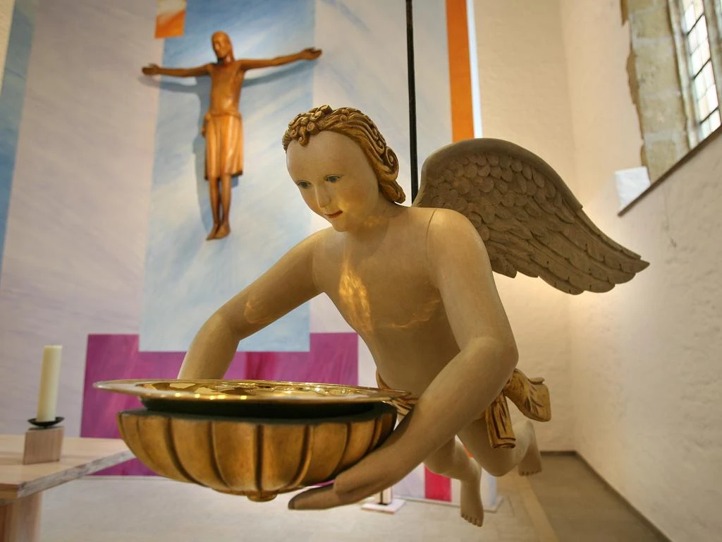 Ein Taufengel schwebt im Kirchenraum, hält eine Schale; im Hintergrund ein Kruzifix zu sehen.
