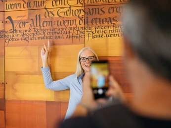 Frau zeigt auf Wand mit Text der Benediktsregel, Besucher fotografiert sie im Abtei Marienmünster Zentrum.