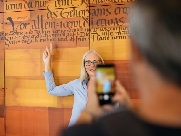 Frau zeigt auf Wand mit Text der Benediktsregel, Besucher fotografiert sie im Abtei Marienmünster Zentrum.
