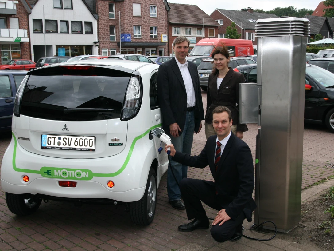 Drei Personen präsentieren ein Elektroauto an einer Ladestation in einer urbanen Umgebung.