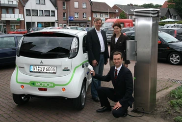 Ladesäule für Elektroautos und -roller Drei Personen präsentieren ein Elektroauto an einer Ladestation in einer urbanen Umgebung.