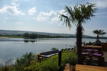 Freizeitanlage Godelheim mit Strandbar Palmen und Holzmöbel an einem Seeufer mit bewölktem Himmel und einer Liegewiese im Hintergrund.