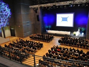 Menschen sitzen in einem modernen, großen Konferenzsaal und verfolgen eine Präsentation auf einer Bühne.