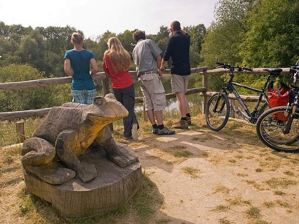 Vier Personen betrachten von einem Geländer aus einen Teich. Ein hölzerner Frosch und Fahrräder sind sichtbar.