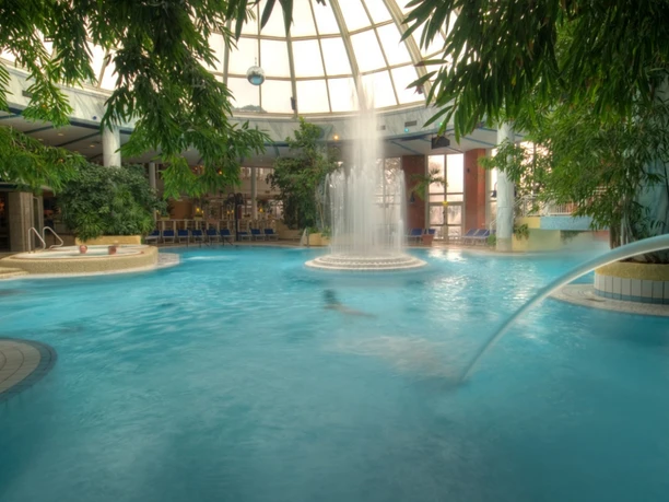 Ein großer Innenpool mit einer Wasserfontäne in der Mitte, umgeben von grüner Bepflanzung.